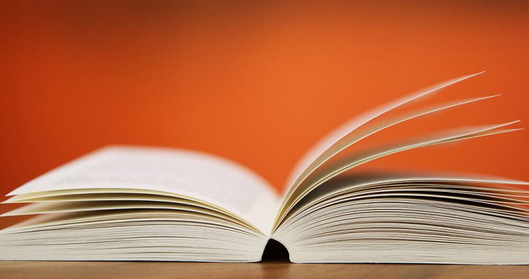 Öppen bok med upplyfta sidor på bord mot orange bakgrund.