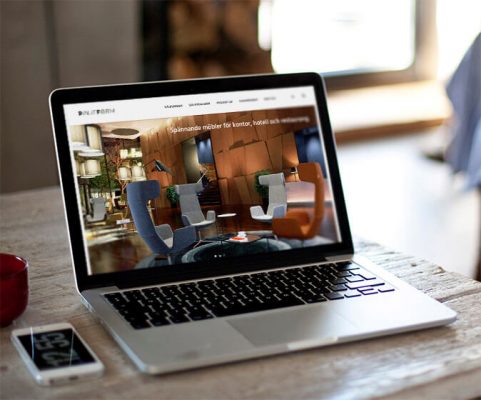 Bärbar dator på bord visar webbplats med moderna kontors- och hotellmöbler, smartphone bredvid.